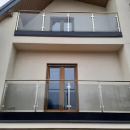 Dwa balkony z balustradami szklanymi na budynku w Nowej Wsi. Balustrady ze stali nierdzewnej z szybami, czarne elementy wykończeniowe.