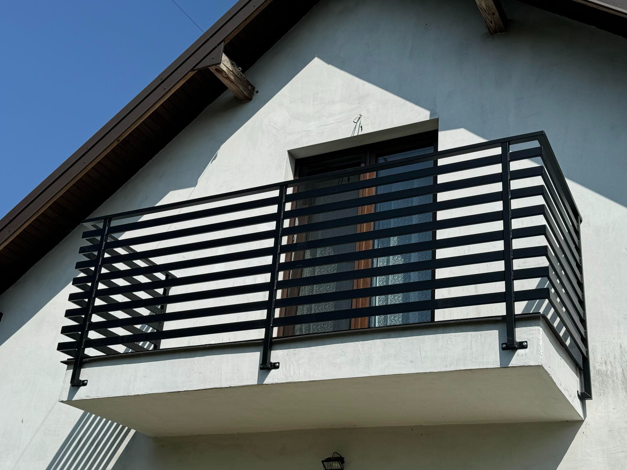 Czarna, pozioma balustrada metalowa na balkonie z widokiem na okno i fragment dachu domu.