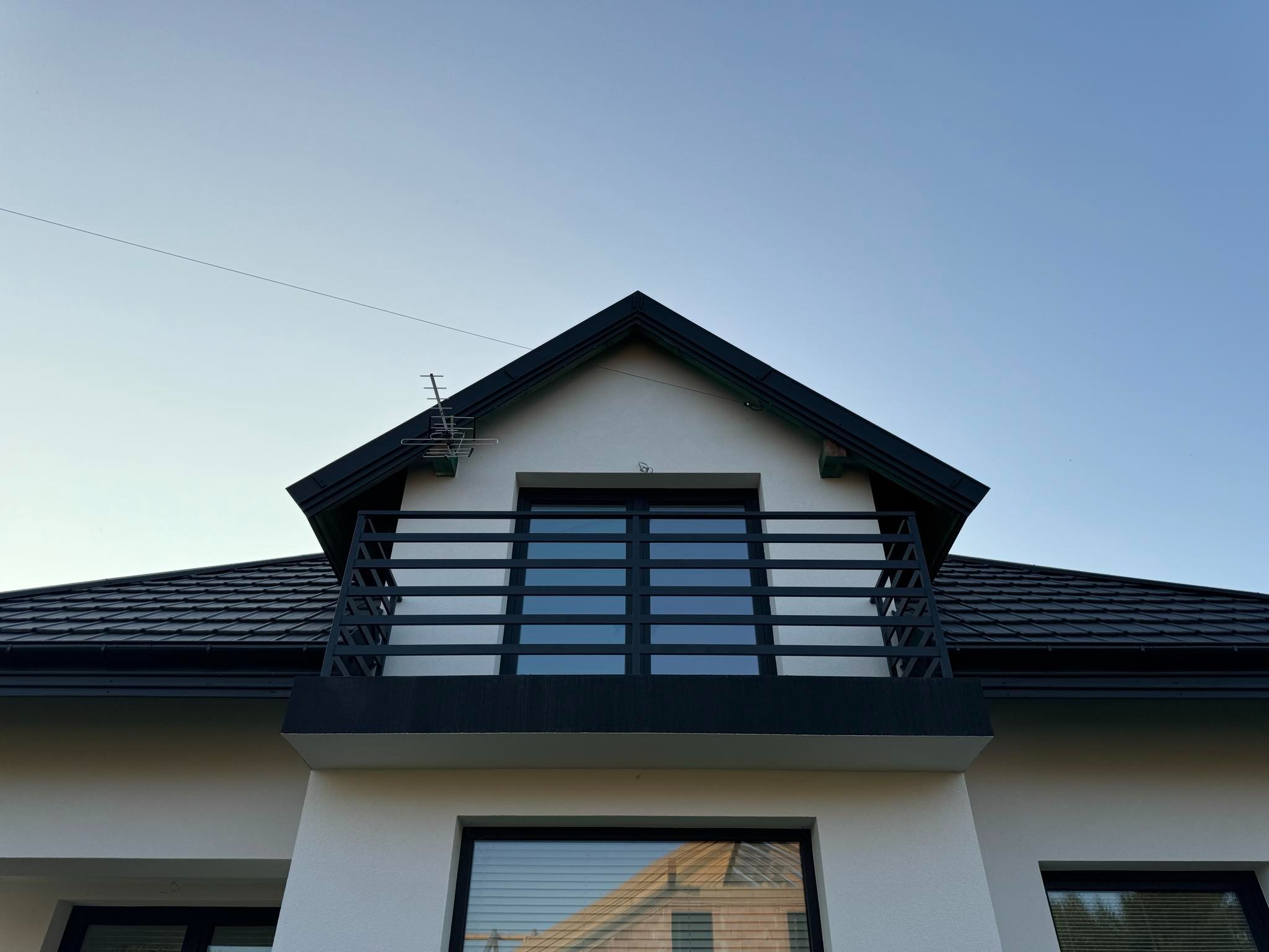 Fragment fasady domu z balkonem zabezpieczonym czarną, metalową konstrukcją o poziomym układzie szczebli, widoczny dach z czarnej blachy oraz okna z czarnymi ramami, na dachu antena telewizyjna.