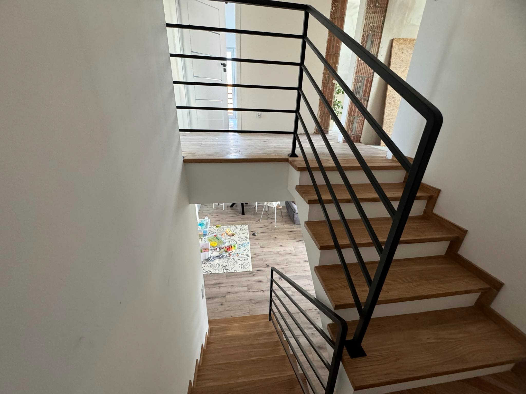 Wewnętrzne schody drewniane z czarną, prostą balustradą z poziomych prętów, widok z góry na dół, z widocznym salonem z dywanem i zabawkami na dole.