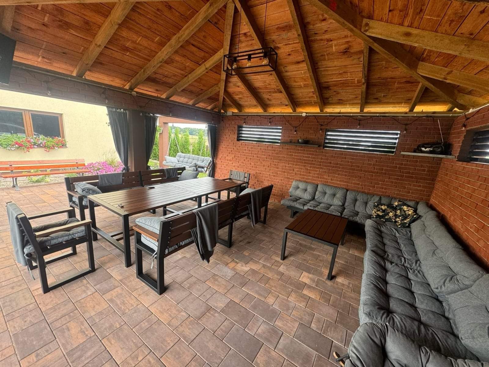 Zadaszona przestrzeń wypoczynkowa z ceglaną ścianą, drewnianym sufitem i kompletem mebli ogrodowych z metalowymi elementami, w tle huśtawka ogrodowa i rośliny.