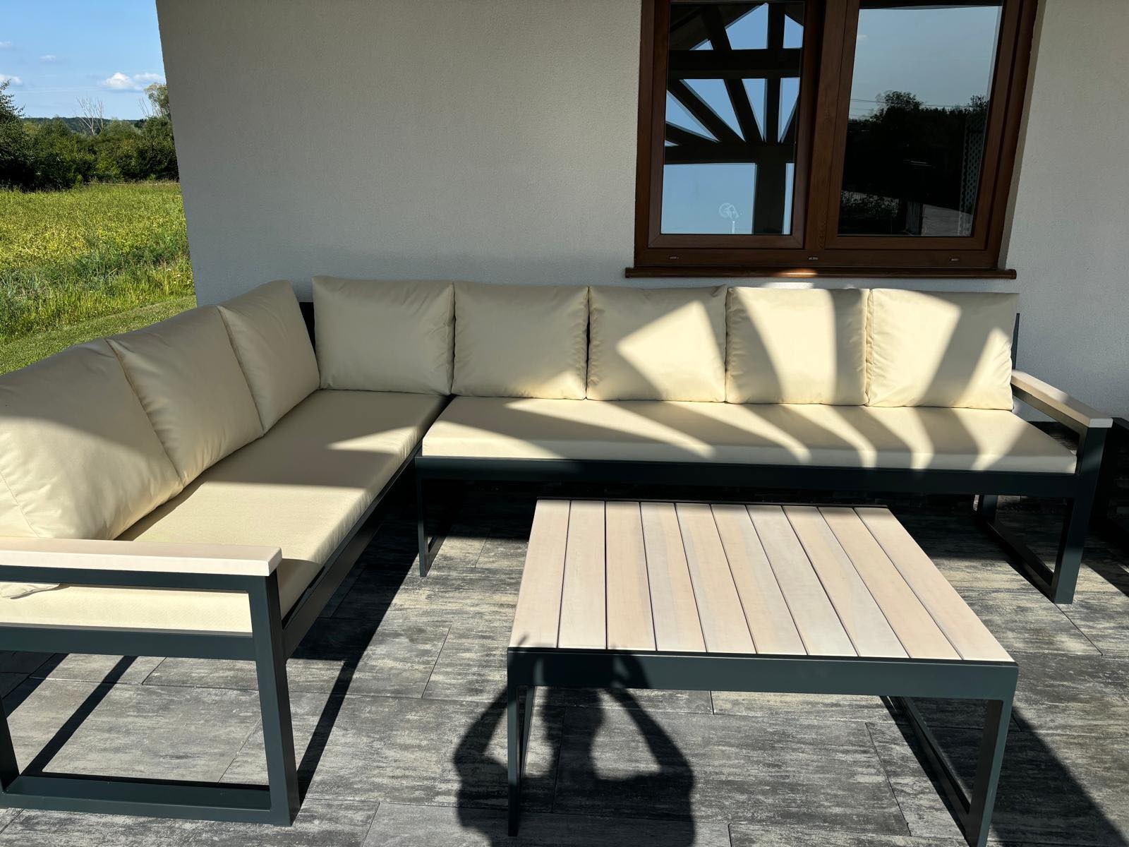 Zestaw mebli ogrodowych: narożna sofa z jasnymi poduszkami i stolik kawowy z drewnianym blatem, na tle białej ściany budynku z oknem i zielenią.