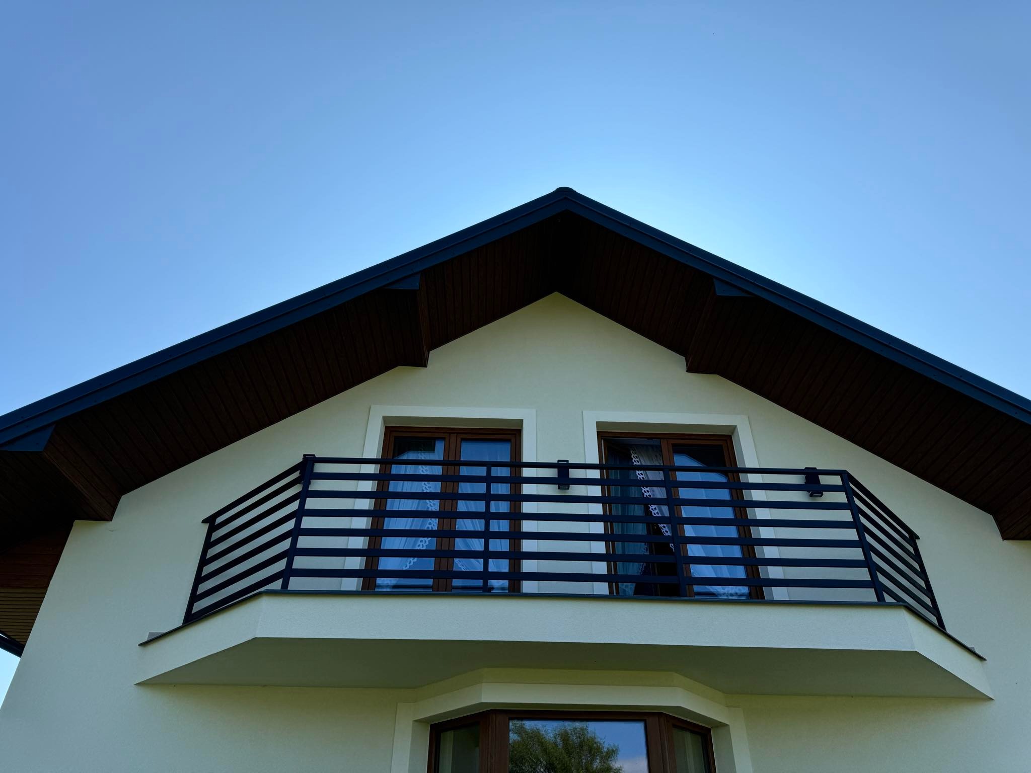 Czarna, pozioma balustrada balkonowa z metalowych profili na tle białej elewacji domu z brązowym dachem i błękitnego nieba.