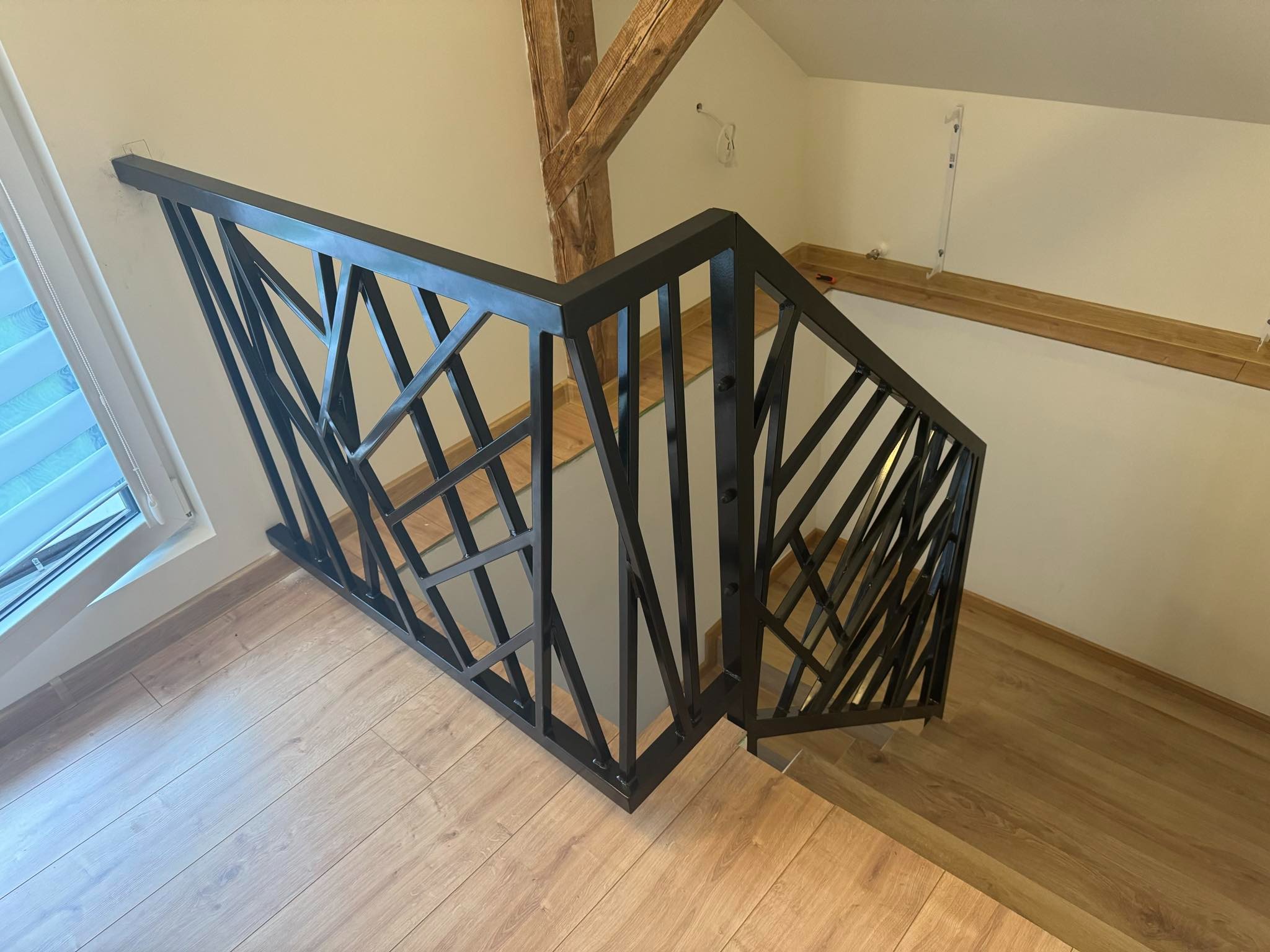 Czarna, geometryczna balustrada schodowa z metalu na drewnianych schodach, widok z góry, z elementami drewnianej konstrukcji stropu widocznymi w tle.