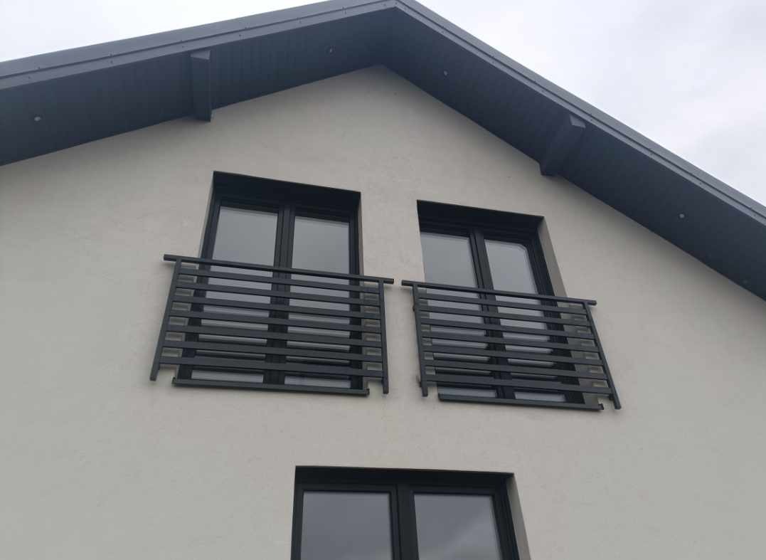 Dwa małe balkony z czarnymi, poziomymi barierkami na tle białej fasady domu z szarym dachem. Trzecie okno poniżej bez balkonu.