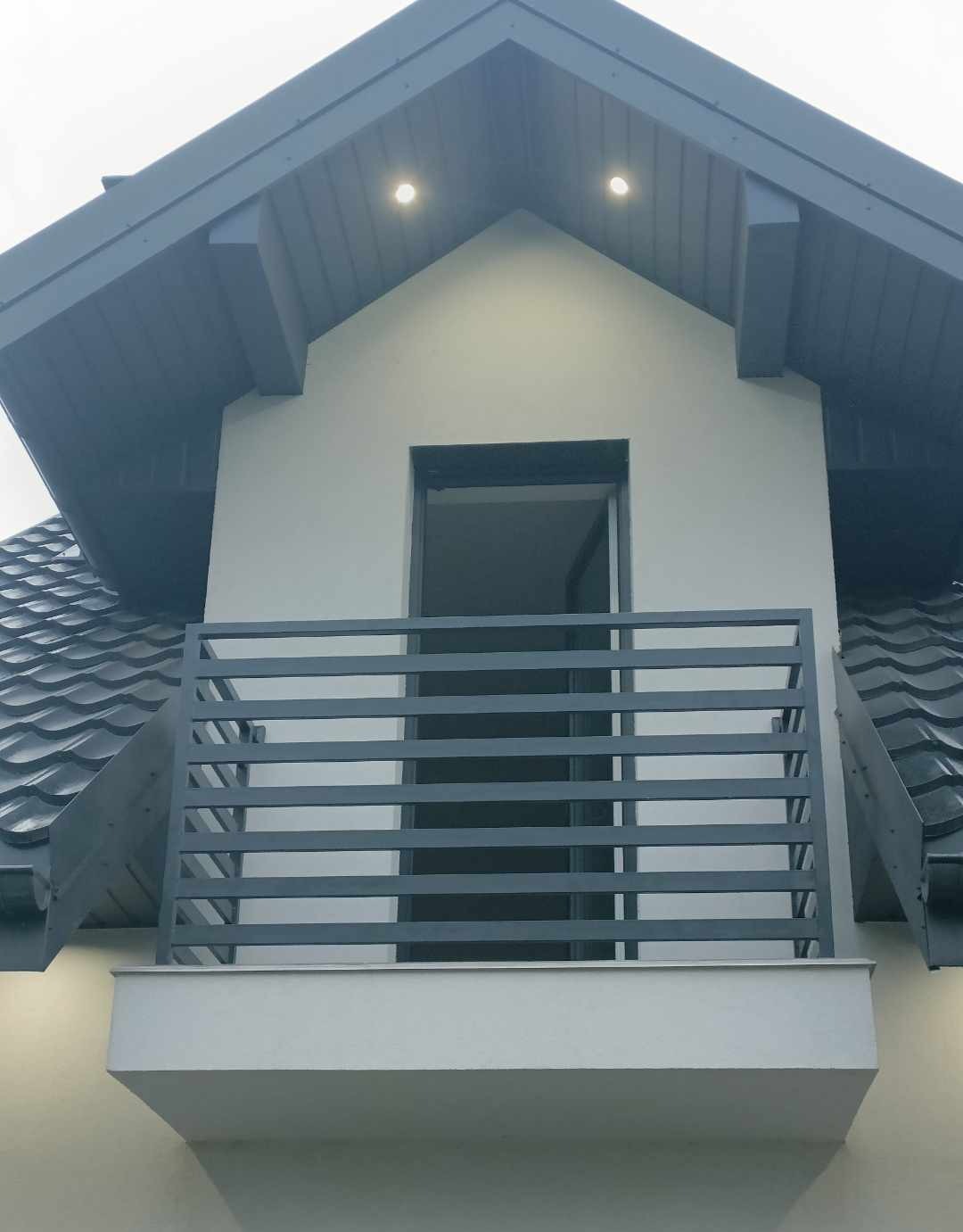 Szara, pozioma balustrada metalowa balkonu na tle białej ściany domu z ciemnoszarym dachem. Widoczne dwa punkty świetlne pod dachem.