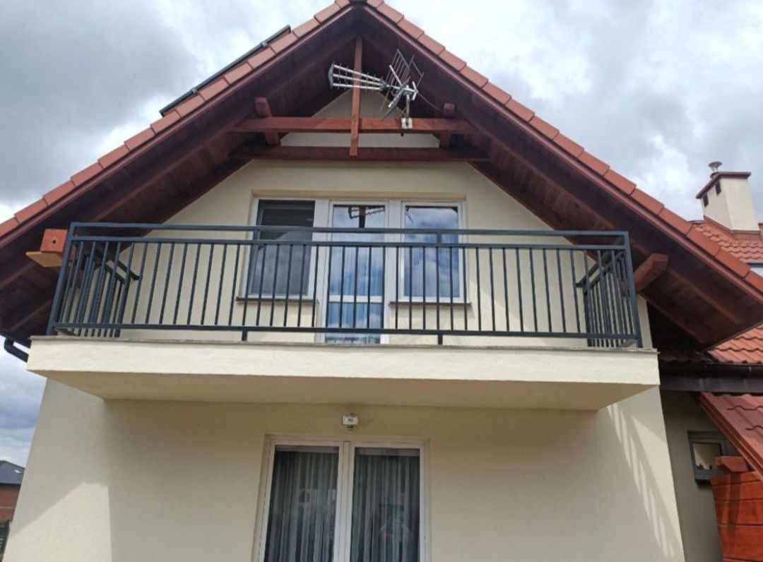 Czarna, metalowa balustrada balkonowa z pionowymi szczeblami na tle kremowej elewacji domu z brązowym dachem i anteną telewizyjną.