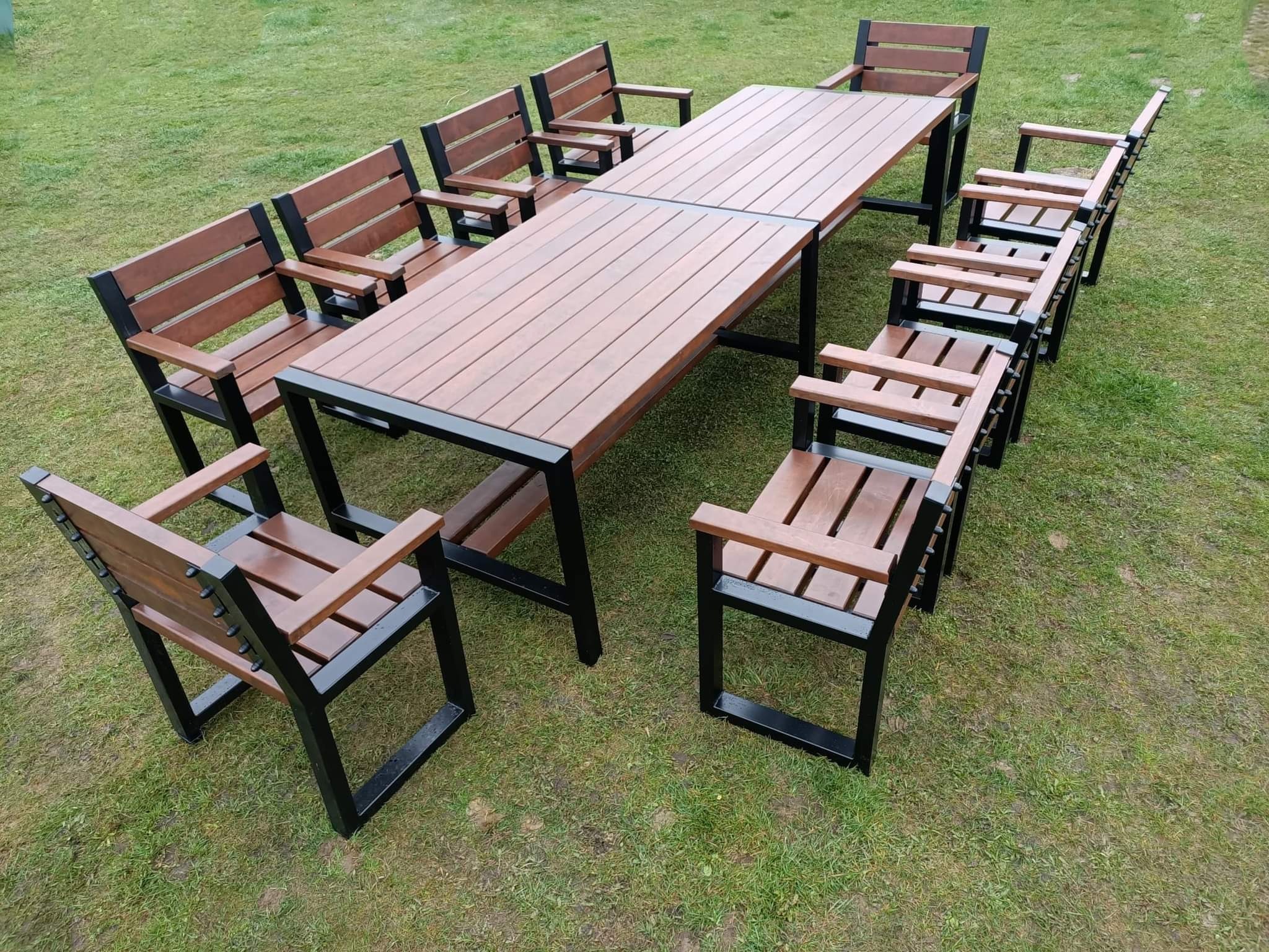 Zestaw mebli ogrodowych: długi stół z drewnianym blatem i metalową ramą oraz dopasowane ławki i krzesła z drewnianymi siedziskami i oparciami, również z metalowymi ramami, ustawione na trawniku.