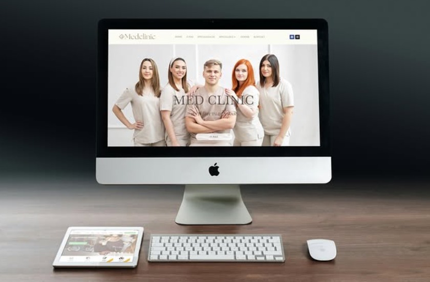 Ekran iMac prezentujący stronę internetową Med Clinic z zespołem medycznym. Tablet, klawiatura i mysz na drewnianym biurku. Ciemne tło.
