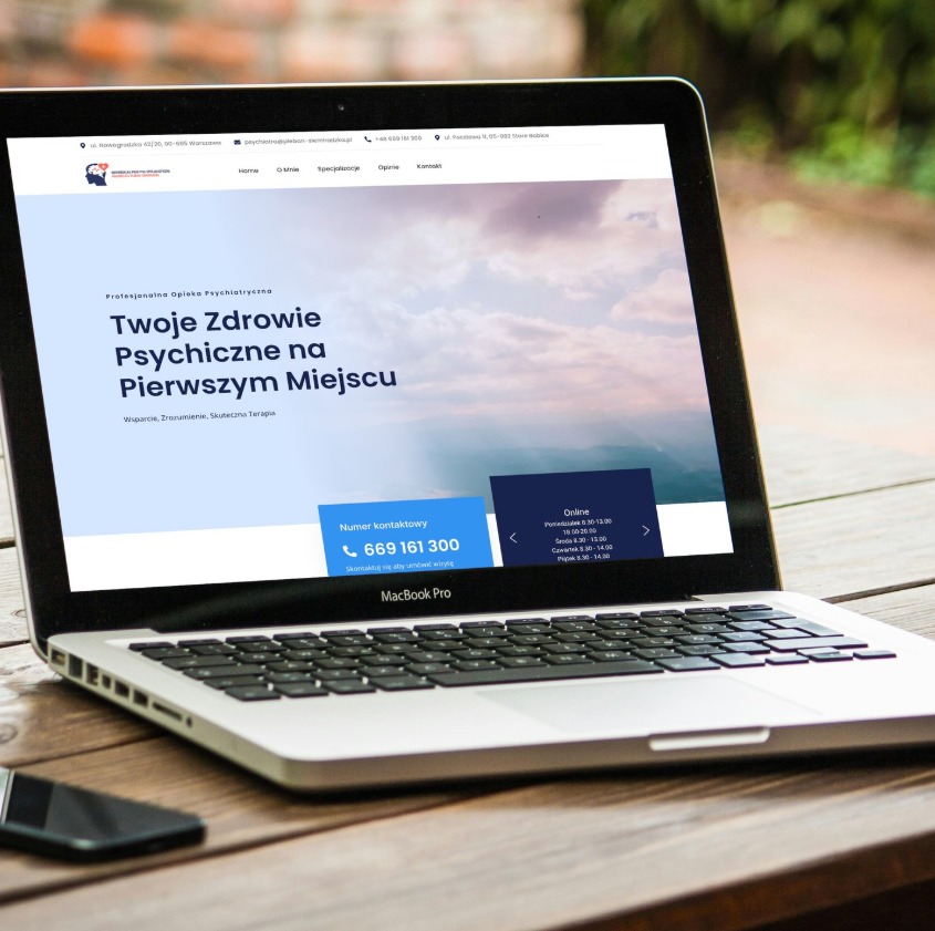 Laptop MacBook Pro na drewnianym stole prezentuje stronę internetową z ofertą psychiatryczną. Widoczny smartfon obok. Jasne tło, naturalne światło.