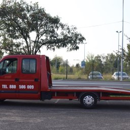 Pogoda w domu - Czerwona laweta Renault Master z numerem telefonu 880 506 009 zaparkowana na asfaltowym placu, widok z boku na tle drzew i dwóch zaparkowanych samochodów osobowych.