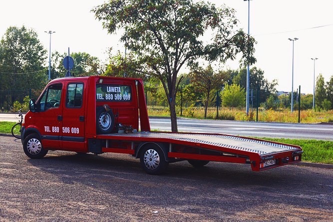 Czerwony pojazd do transportu pojazdów z kołem zapasowym i numerem telefonu na karoserii, zaparkowany na parkingu w słoneczny dzień.