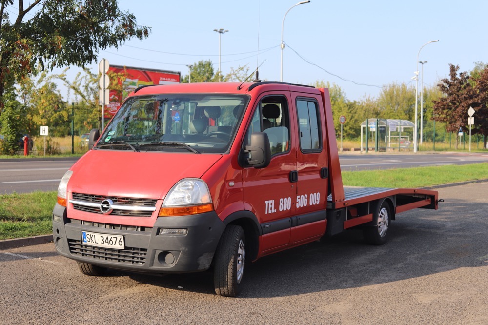 Czerwony samochód transportowy Opel Master z platformą załadunkową stoi na parkingu, widoczny numer telefonu na karoserii, w tle drzewa i przystanek autobusowy.