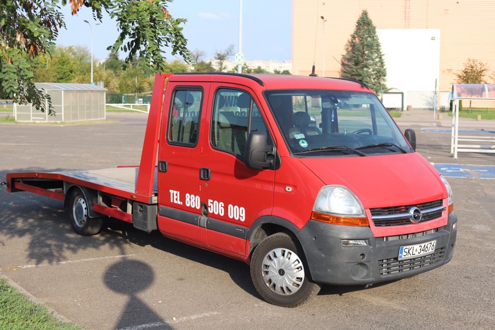 Czerwony samochód z platformą transportową marki Opel, zaparkowany na parkingu, z numerem telefonu umieszczonym na drzwiach.
