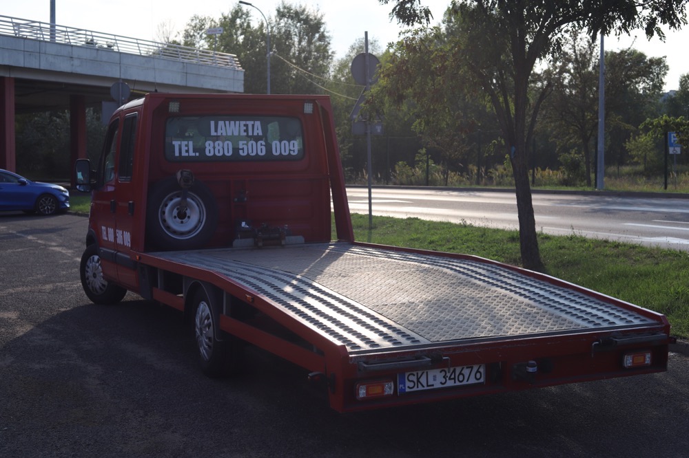 Czerwona platforma transportowa z numerem telefonu na szybie, zaparkowana obok drogi pod wiaduktem.