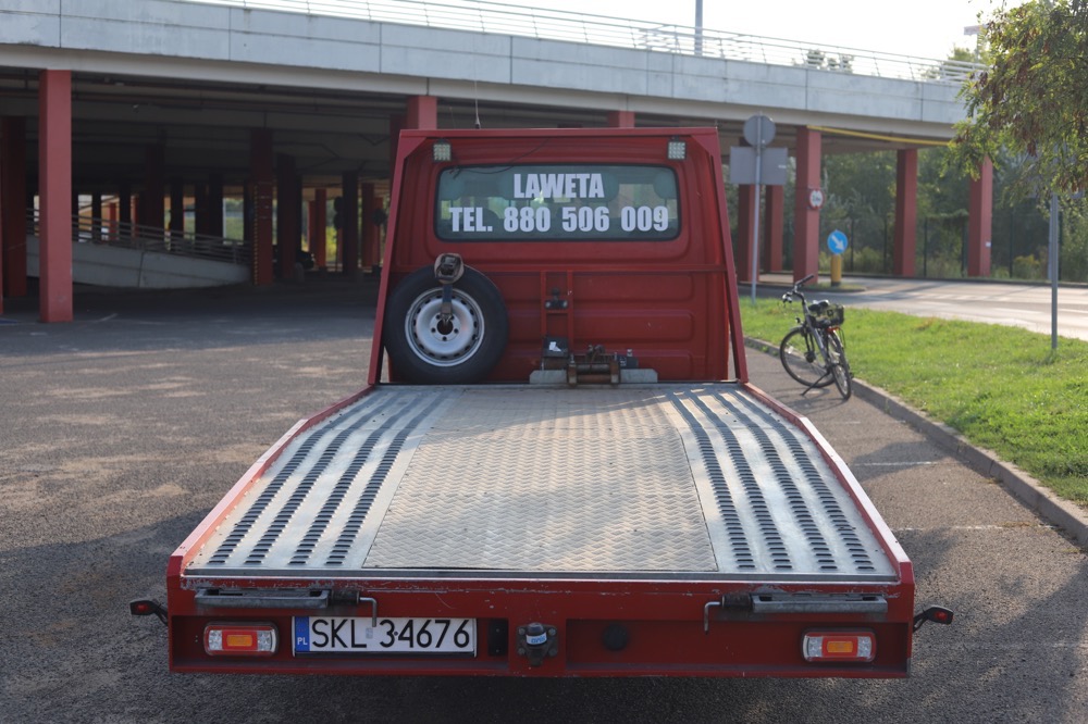 Czerwona platforma transportowa z widocznym numerem telefonu na tylnej szybie, zaparkowana na parkingu z widokiem na betonową konstrukcję i rower w tle.