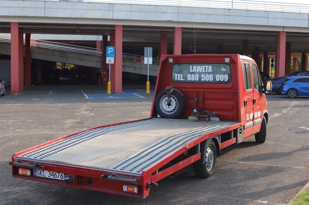 Czerwony pojazd do transportu samochodów z kołem zapasowym z tyłu, zaparkowany na parkingu z widocznym znakiem parkingowym. Na tylnej szybie numer telefonu.