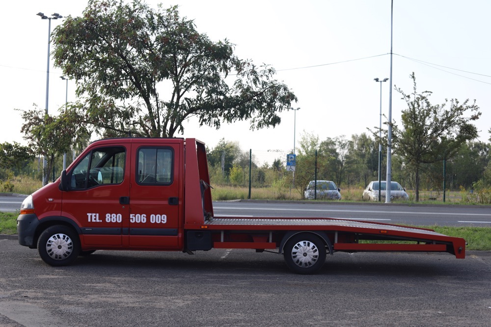 Czerwona laweta Renault Master z numerem telefonu 880 506 009 zaparkowana na asfaltowym placu, widok z boku na tle drzew i dwóch zaparkowanych samochodów osobowych.