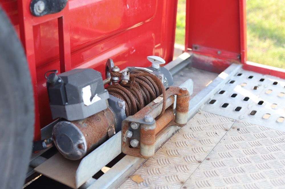 Widok z bliska na rdzewiejącą wciągarkę z liną stalową zamontowaną na platformie pojazdu transportowego, z widocznymi śladami użytkowania i czerwonym elementem nadwozia w tle.