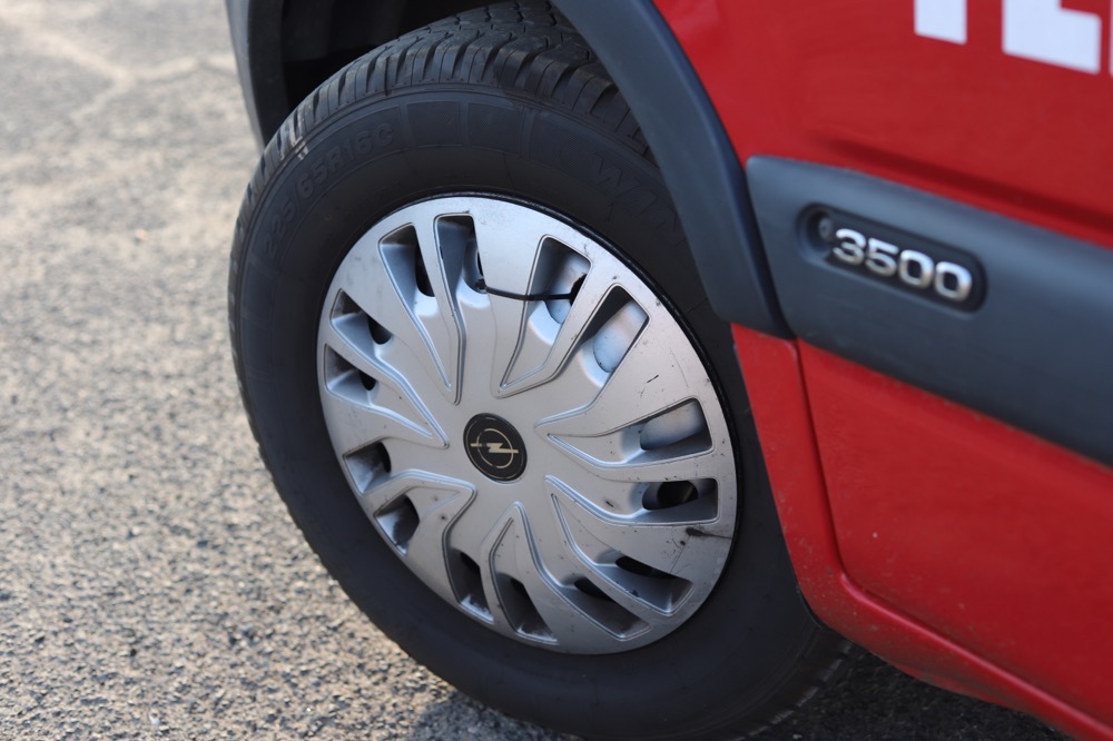 Koło samochodu z felgą Opel, widoczne z boku, na tle asfaltowej nawierzchni w Warszawie. Czerwony lakier karoserii i oznaczenie '3500' na boku pojazdu.