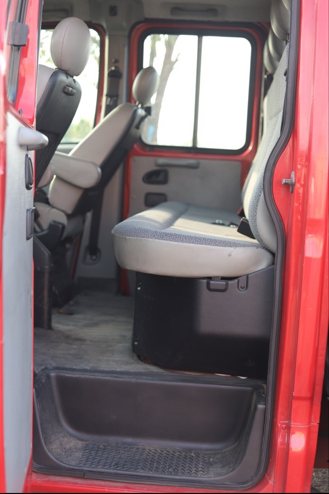 Wnętrze kabiny czerwonego pojazdu transportowego z widocznymi fotelami, pasami bezpieczeństwa i oknem z jasnym tłem.