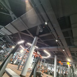 Climat Development - Widok na sufit w siłowni z widocznymi elementami wentylacji, kanałami instalacyjnymi, lampami i kolumnami, z perspektywy żabiej.