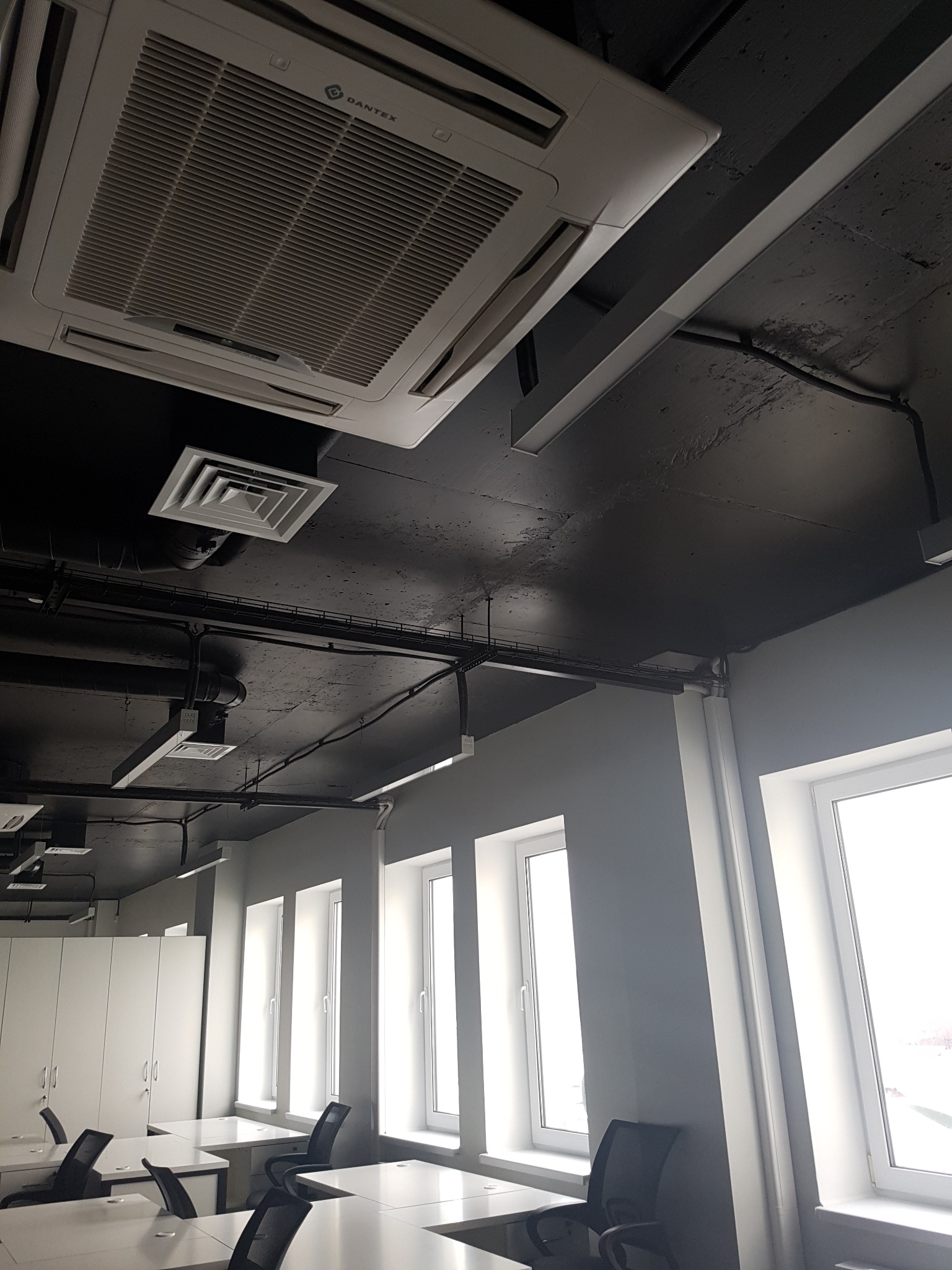 Sufitowe urządzenie klimatyzacyjne marki Dantex w biurze z czarnym sufitem i rzędem wysokich okien.