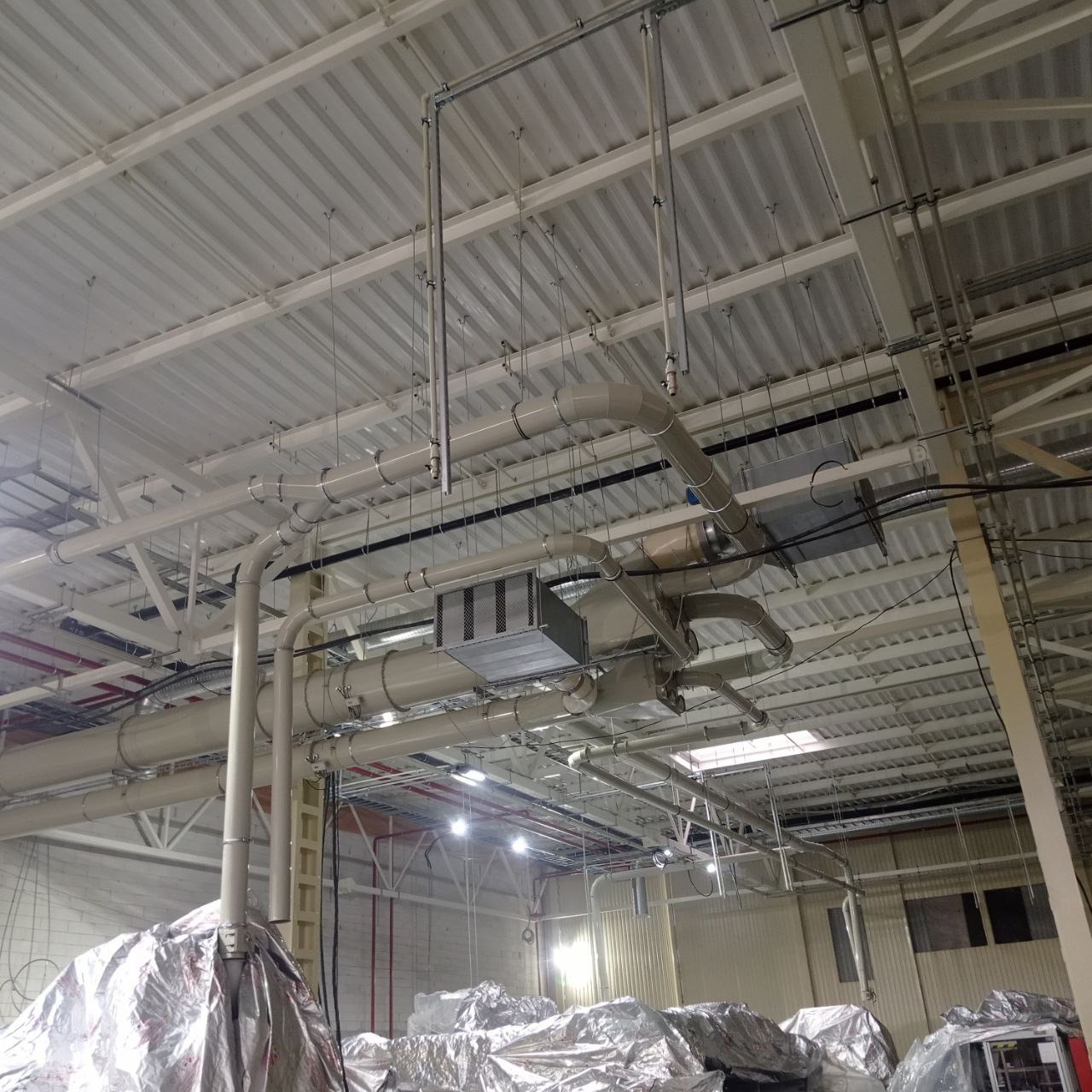 Rozbudowany system wentylacyjny z rurami i kratkami wentylacyjnymi pod sufitem hali przemysłowej, z widocznymi elementami izolacji termicznej na podłodze.