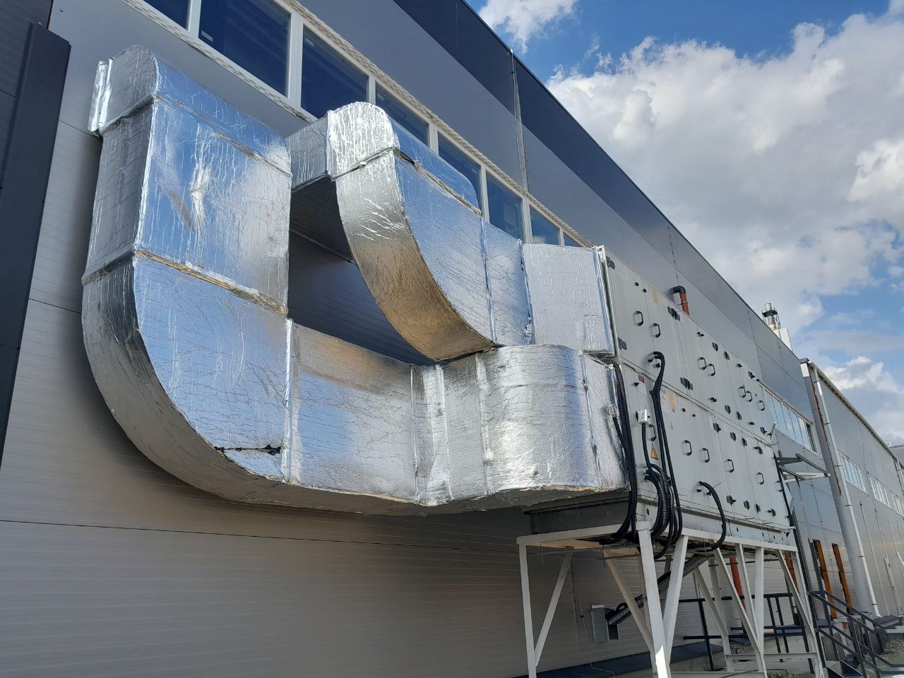 Zewnętrzny montaż izolowanych kanałów wentylacyjnych na ścianie budynku przemysłowego, widoczne elementy instalacji na metalowej konstrukcji wsporczej.
