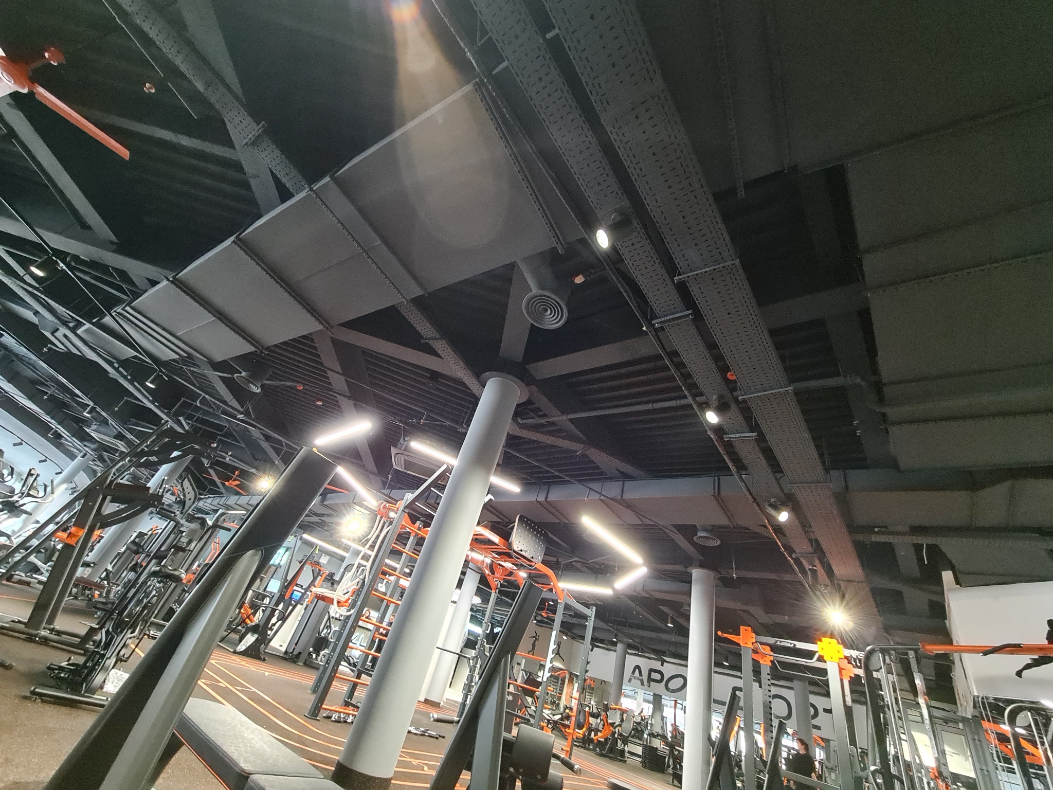 Widok na sufit w siłowni z widocznymi elementami wentylacji, kanałami instalacyjnymi, lampami i kolumnami, z perspektywy żabiej.