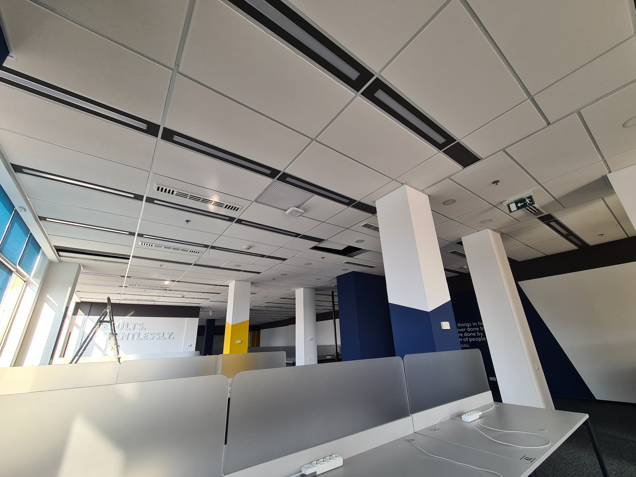 Sufit podwieszany z panelami rastrowymi i liniowymi oprawami oświetleniowymi w nowoczesnym biurze, widoczne kratki wentylacyjne i fragmentarycznie biurka z przegrodami.