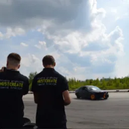 Dwóch mężczyzn w czarnych koszulkach z napisem 'STUDIO GRAFICZNE MUSTCREATE Dawid Sroczyk' obserwuje czarny samochód na torze wyścigowym pod błękitnym niebem z chmurami.