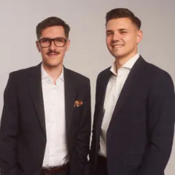 Portret dwóch uśmiechniętych mężczyzn w garniturach na jasnym tle, jeden z wąsami i okularami, drugi z krótką fryzurą.