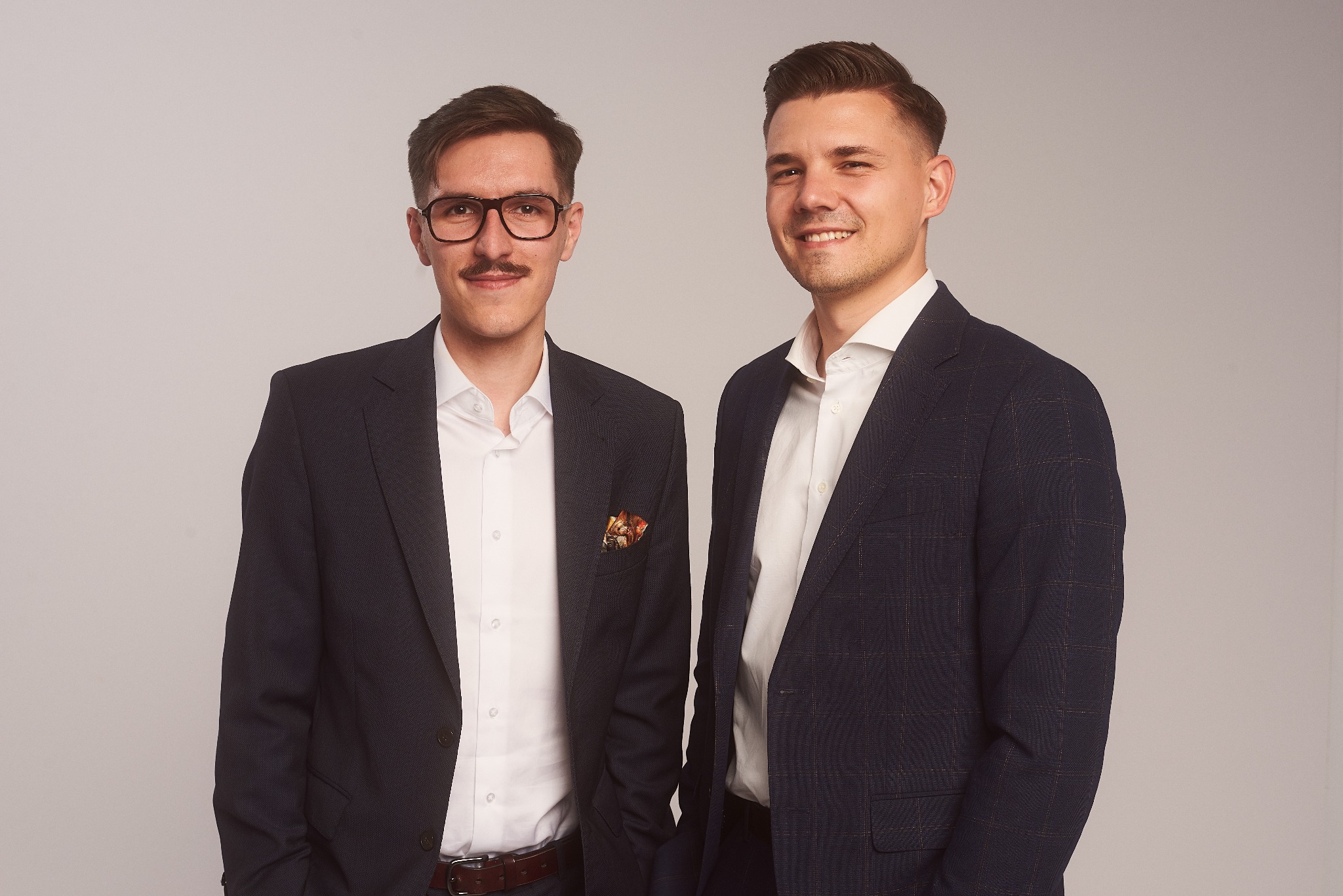 Portret dwóch uśmiechniętych mężczyzn w garniturach na jasnym tle, jeden z wąsami i okularami, drugi z krótką fryzurą.
