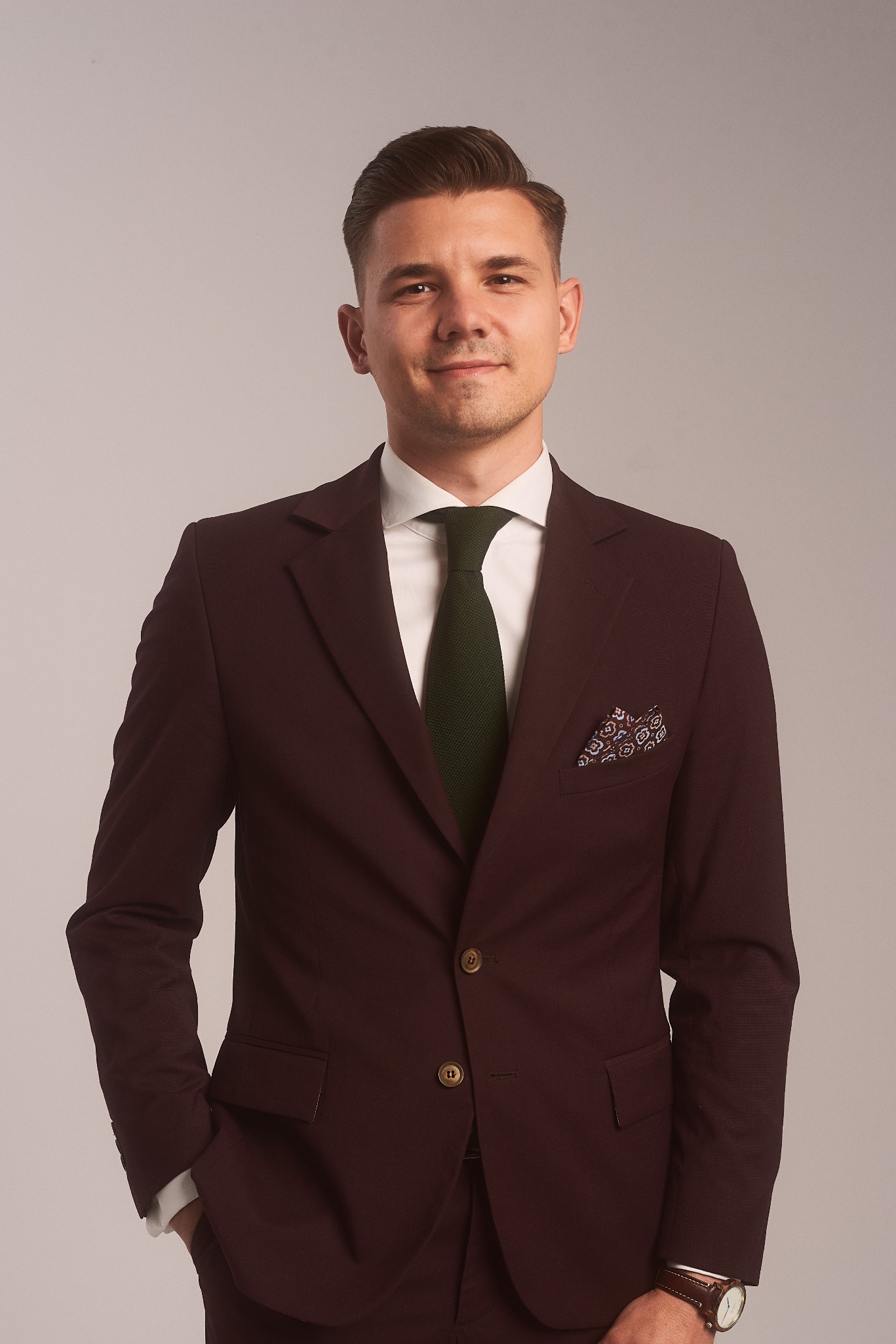 Portret mężczyzny w bordowym garniturze, białej koszuli i zielonym krawacie, z ozdobną poszetką w kieszonce, pozującego na neutralnym tle.
