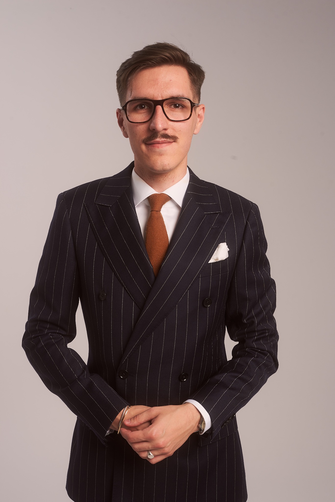 Portret młodego adwokata w dwurzędowym garniturze w prążki, z brązowym krawatem i okularami, stojącego na jasnoszarym tle.