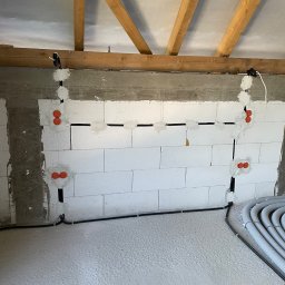 Łukasz Cachro Instalacje elektryczne - Ściana z instalacją elektryczną w trakcie montażu: przewody w bruzdach, puszki pod gniazdka, widoczne rury osłonowe na podłodze. Surowy stan, poddasze z drewnianą konstrukcją.