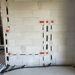 Łukasz Cachro Instalacje elektryczne - Ściana z betonu komórkowego z wyfrezowanymi pionowymi i poziomymi kanałami na przewody elektryczne, zabezpieczonymi pomarańczowymi puszkami. Widoczne kable w podłodze.