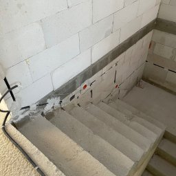 Łukasz Cachro Instalacje elektryczne - Betonowe schody w surowym stanie z widoczną instalacją elektryczną w peszlach, wyprowadzoną ze ściany z betonu komórkowego. Czerwone puszki instalacyjne.