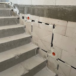 Łukasz Cachro Instalacje elektryczne - Montaż przewodów w ścianie przy schodach, widoczne peszle i kable. Prace instalacyjne w surowym stanie budynku, przed wykończeniem.
