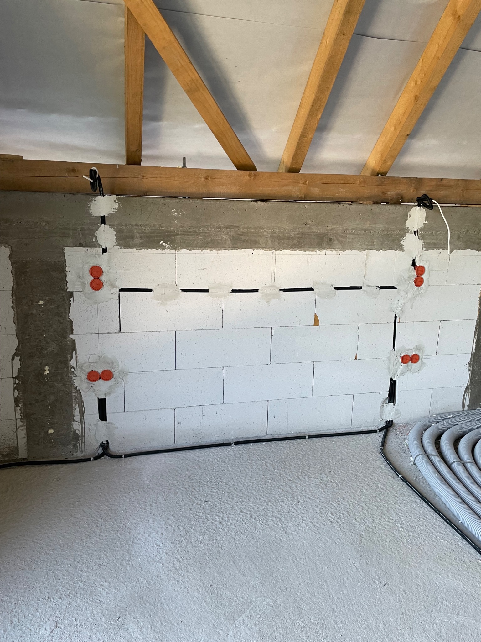 Ściana z instalacją elektryczną w trakcie montażu: przewody w bruzdach, puszki pod gniazdka, widoczne rury osłonowe na podłodze. Surowy stan, poddasze z drewnianą konstrukcją.