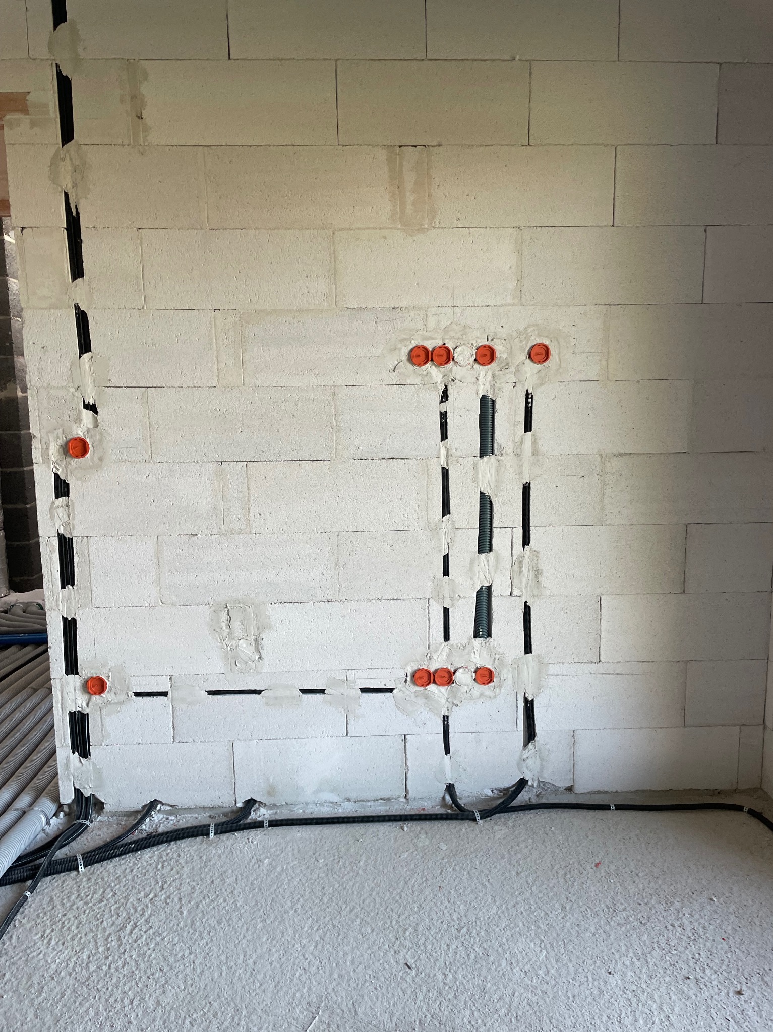 Ściana z betonu komórkowego z wyfrezowanymi pionowymi i poziomymi kanałami na przewody elektryczne, zabezpieczonymi pomarańczowymi puszkami. Widoczne kable w podłodze.