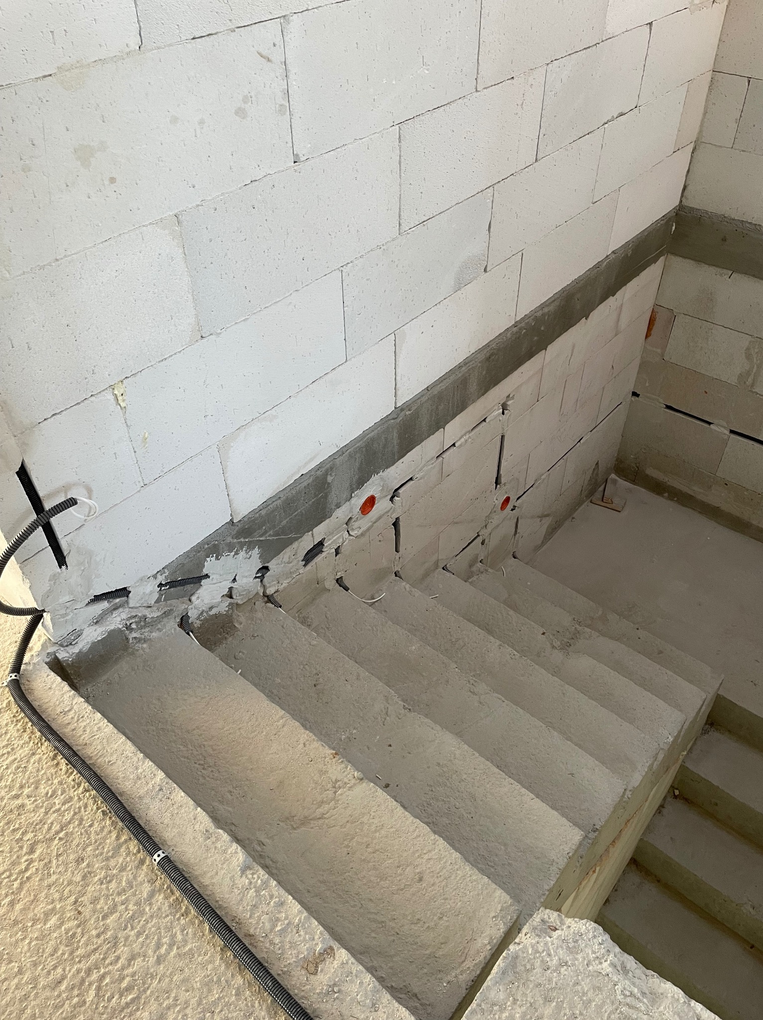 Betonowe schody w surowym stanie z widoczną instalacją elektryczną w peszlach, wyprowadzoną ze ściany z betonu komórkowego. Czerwone puszki instalacyjne.