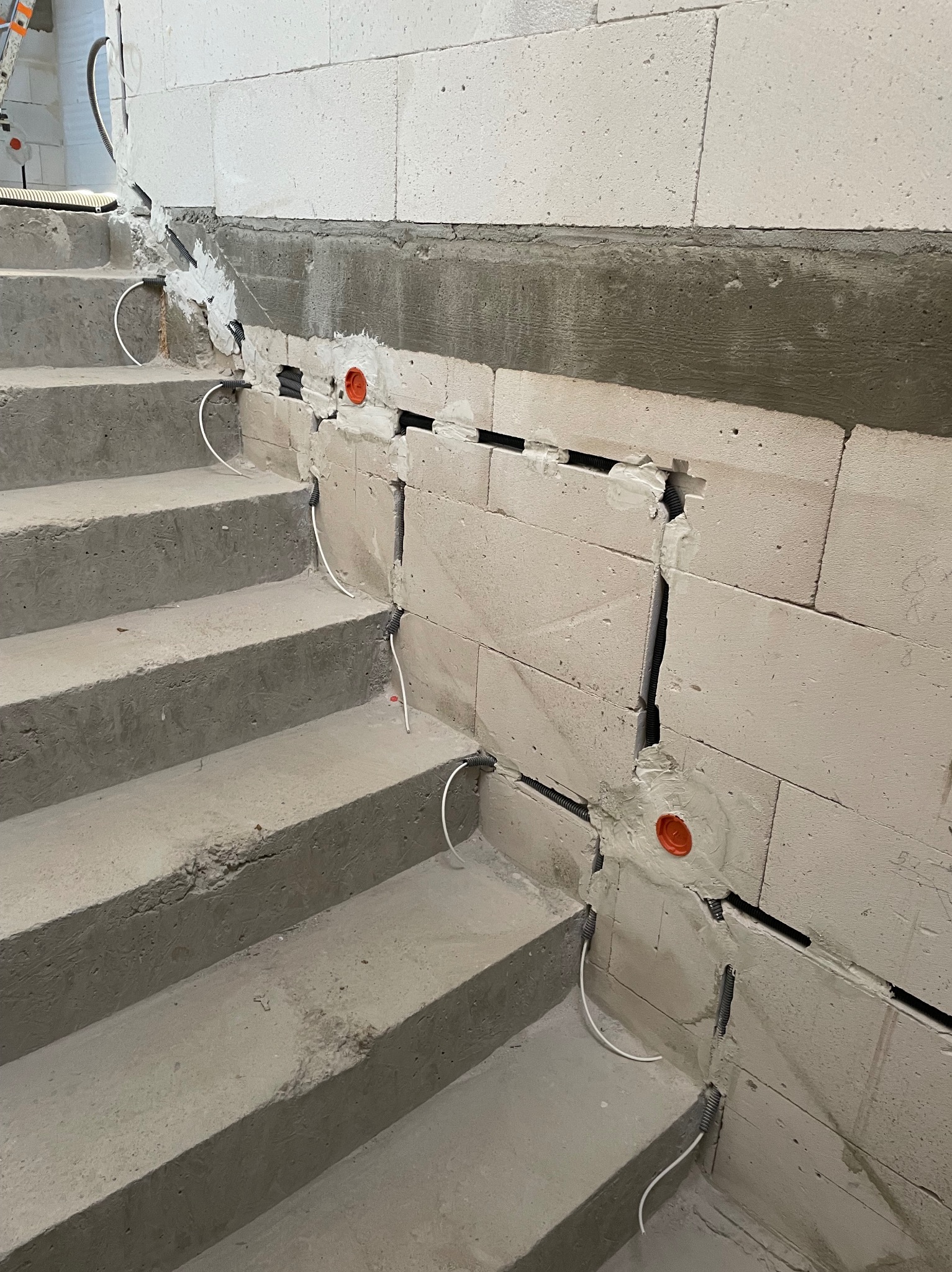 Montaż przewodów w ścianie przy schodach, widoczne peszle i kable. Prace instalacyjne w surowym stanie budynku, przed wykończeniem.
