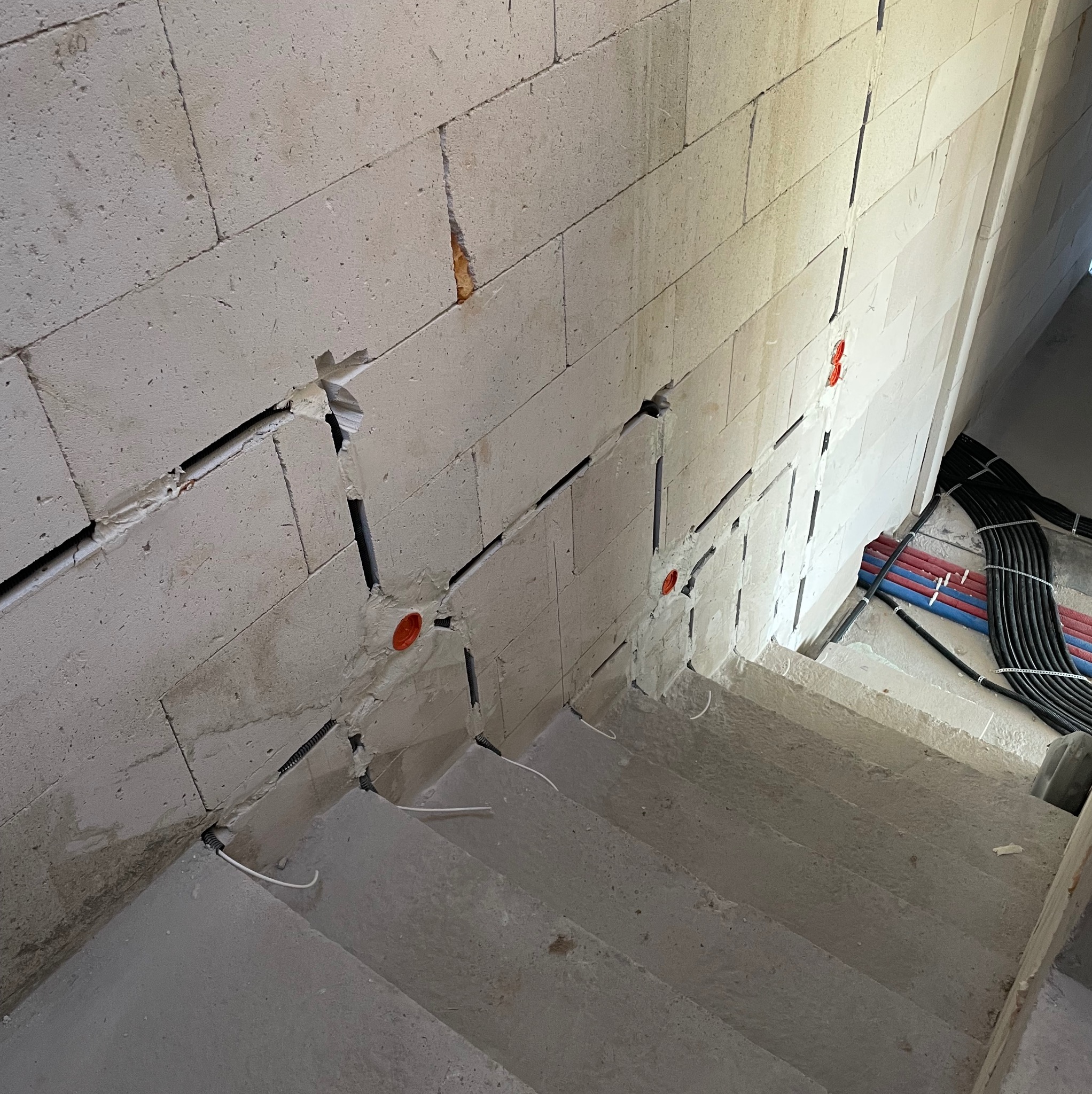 Wykute bruzdy w ścianie z betonu komórkowego pod instalacje elektryczne, widoczne peszle i puszki. W tle rury i przewody. Klatka schodowa w surowym stanie.