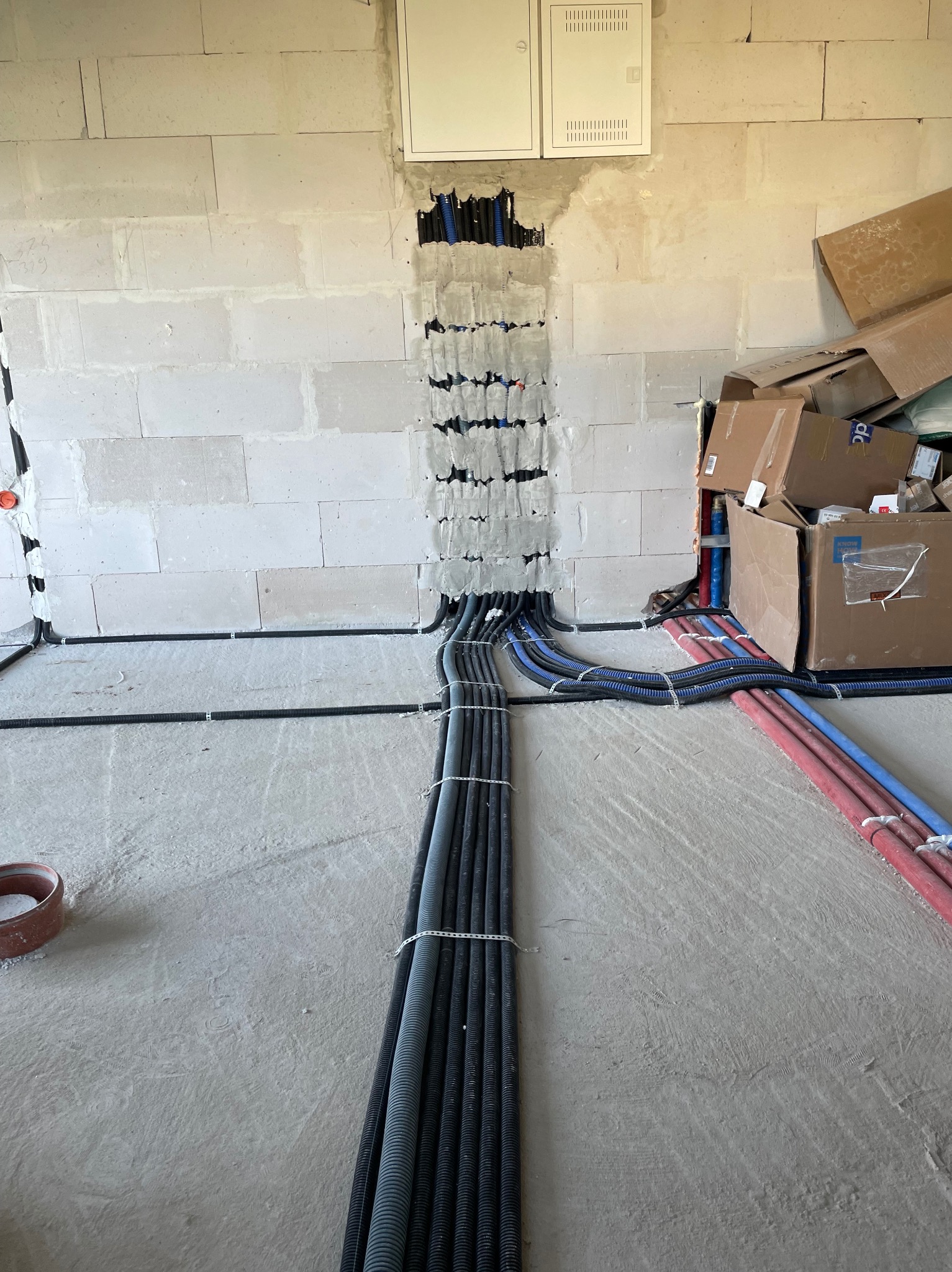 Instalacja przewodów w ścianie z betonu komórkowego. Widoczne kable w peszlach prowadzone do skrzynki elektrycznej. Część kabli ułożona na podłodze.