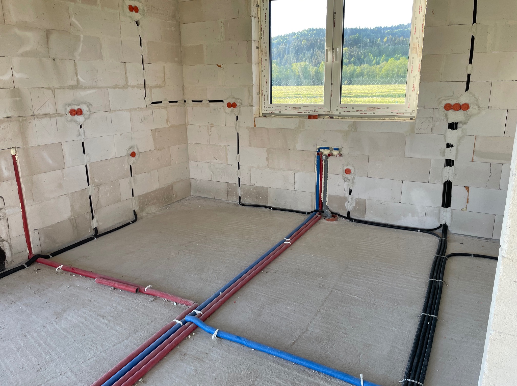 Surowy stan: instalacje elektryczne i hydrauliczne w budynku z pustaków, widoczne przewody w ścianach i rury na podłodze, okno z widokiem na zieleń.