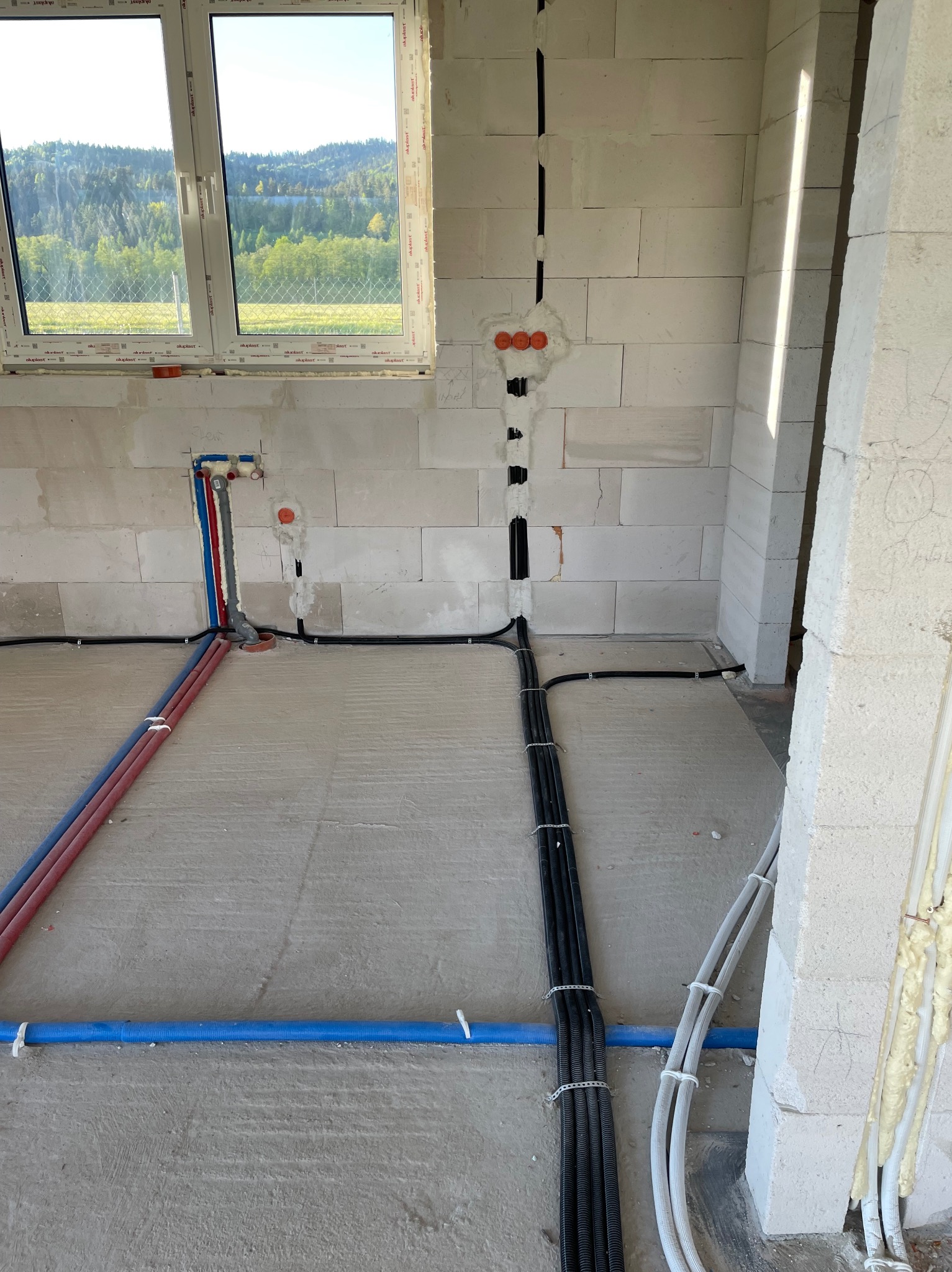 Surowy stan: instalacja elektryczna i wodna w budynku z oknem z widokiem na las. Kable w peszlach, rury w kolorach niebieskim i czerwonym, ściany z białego betonu komórkowego.