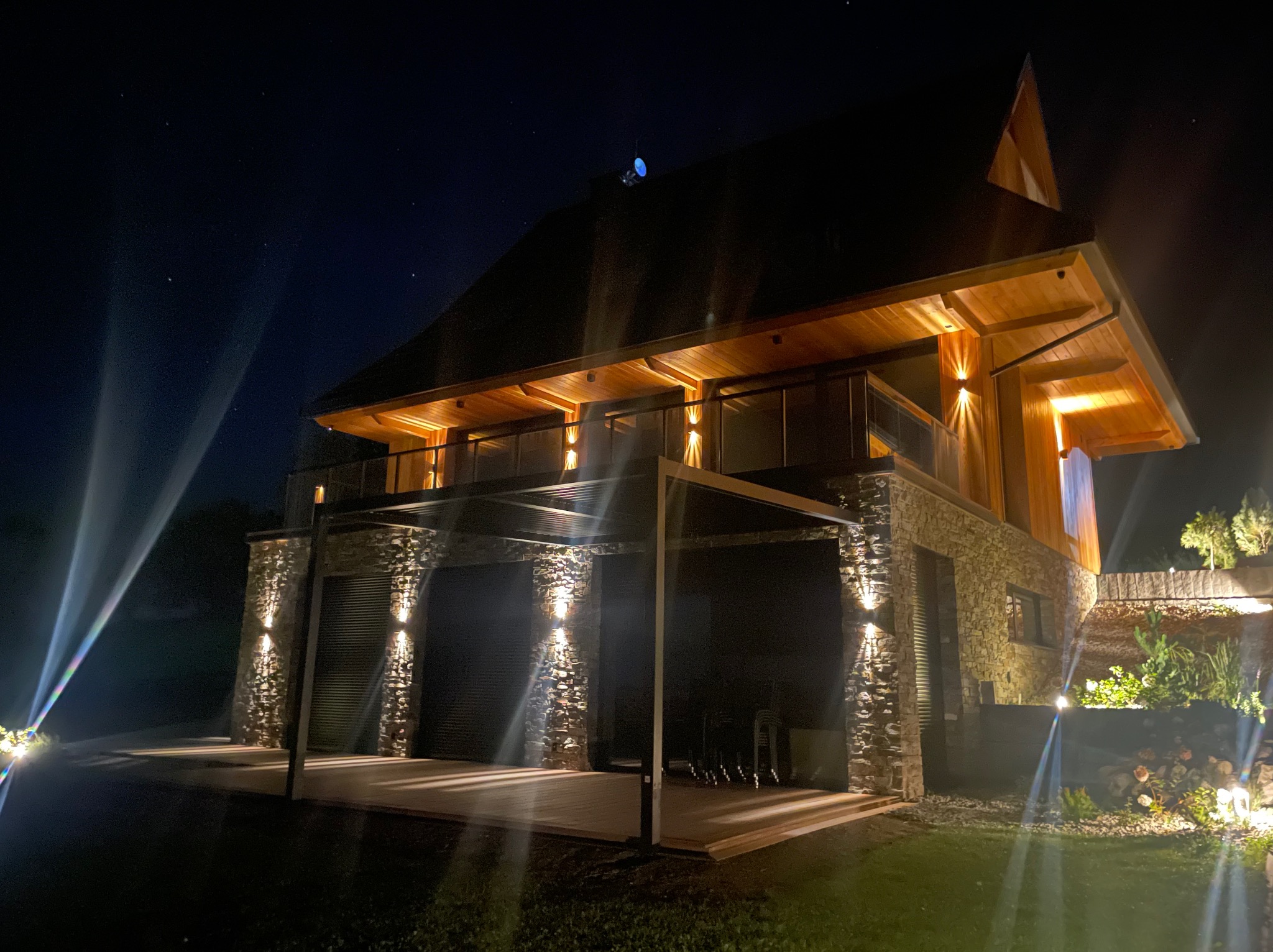 Dwupiętrowy dom z kamienia i drewna nocą, oświetlony ciepłymi lampami ściennymi i reflektorami, podkreślającymi architekturę i detale elewacji.