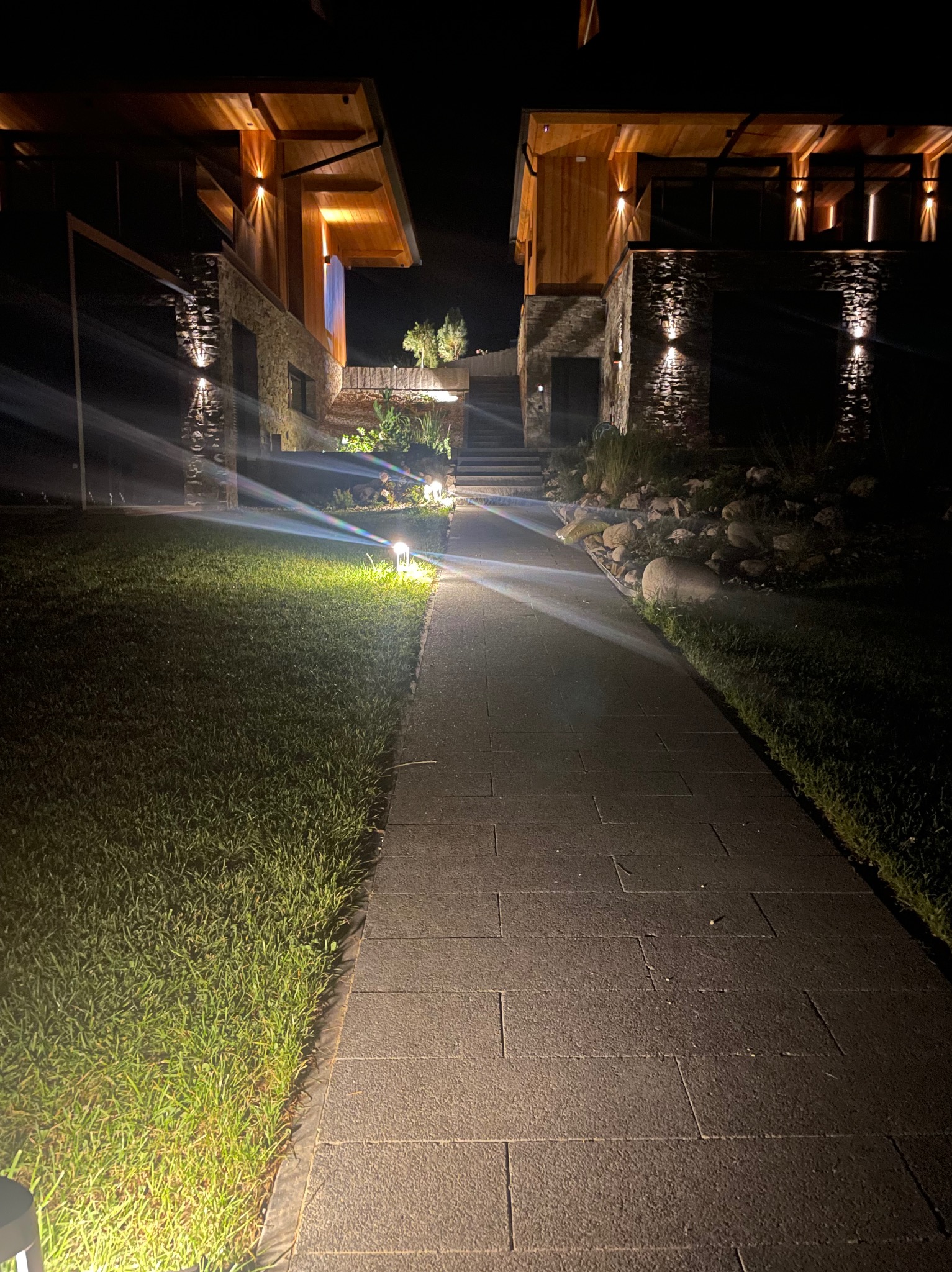 Oświetlona ścieżka prowadząca do dwóch budynków z kamienną elewacją i drewnianymi elementami, nocą. Lampy ogrodowe i elewacyjne tworzą ciepłą atmosferę.